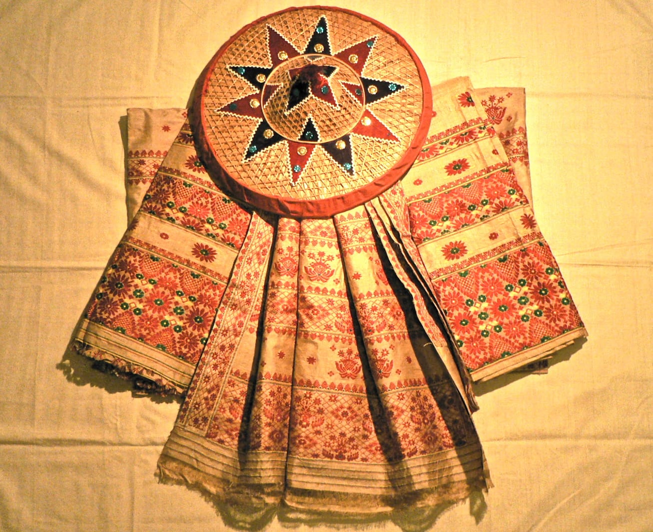 A length of golden muga silk mekhela-chador woven with red and gold floral and pictorial borders, laid out with a decorated japi