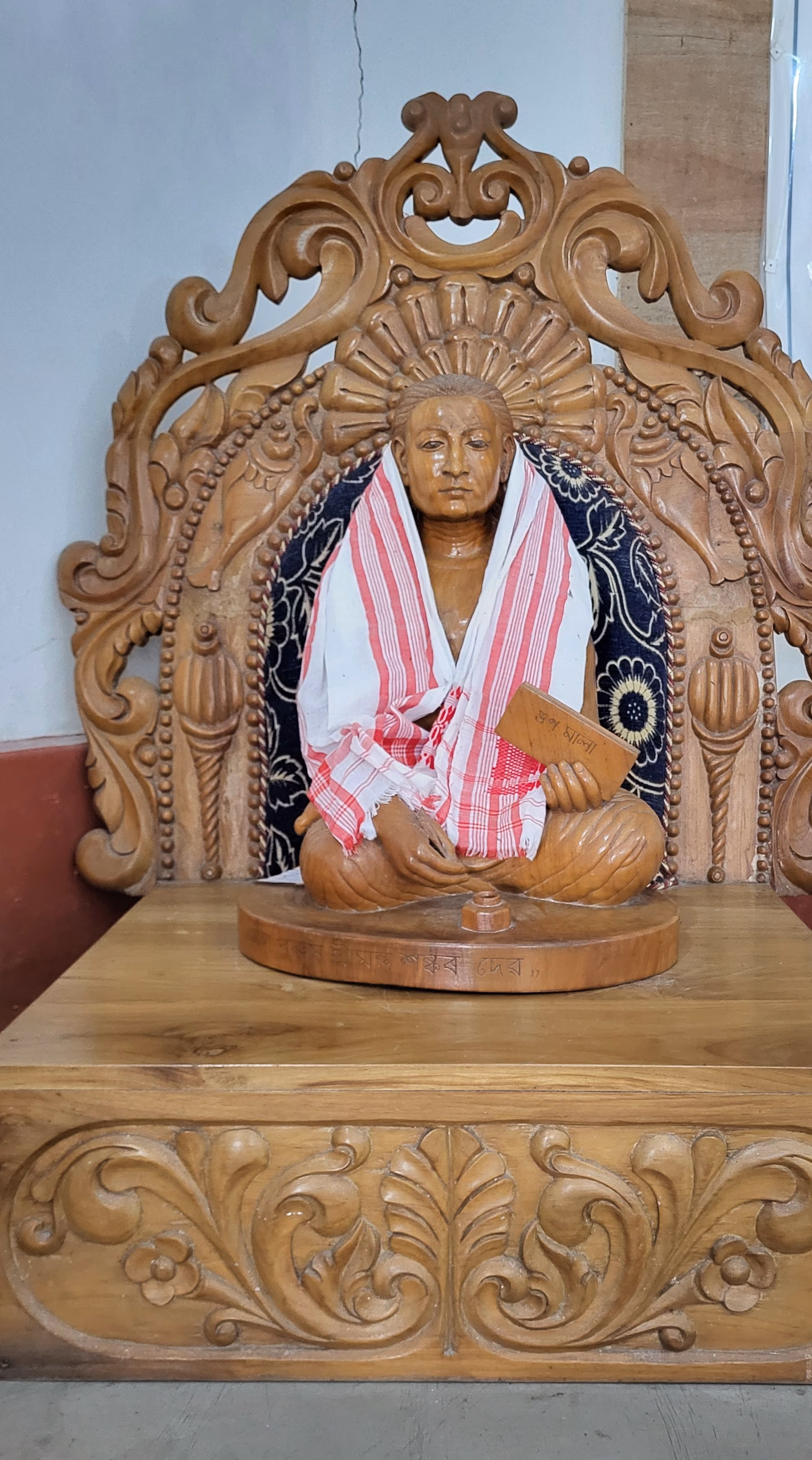 Carved wooden statue of Srimanta Sankardev seated cross-legged within an ornately carved wooden throne with a sunburst halo behind his head, draped in a red-and-white gamosa over a dark floral shawl and holding a small manuscript, on an inscribed carved pedestal