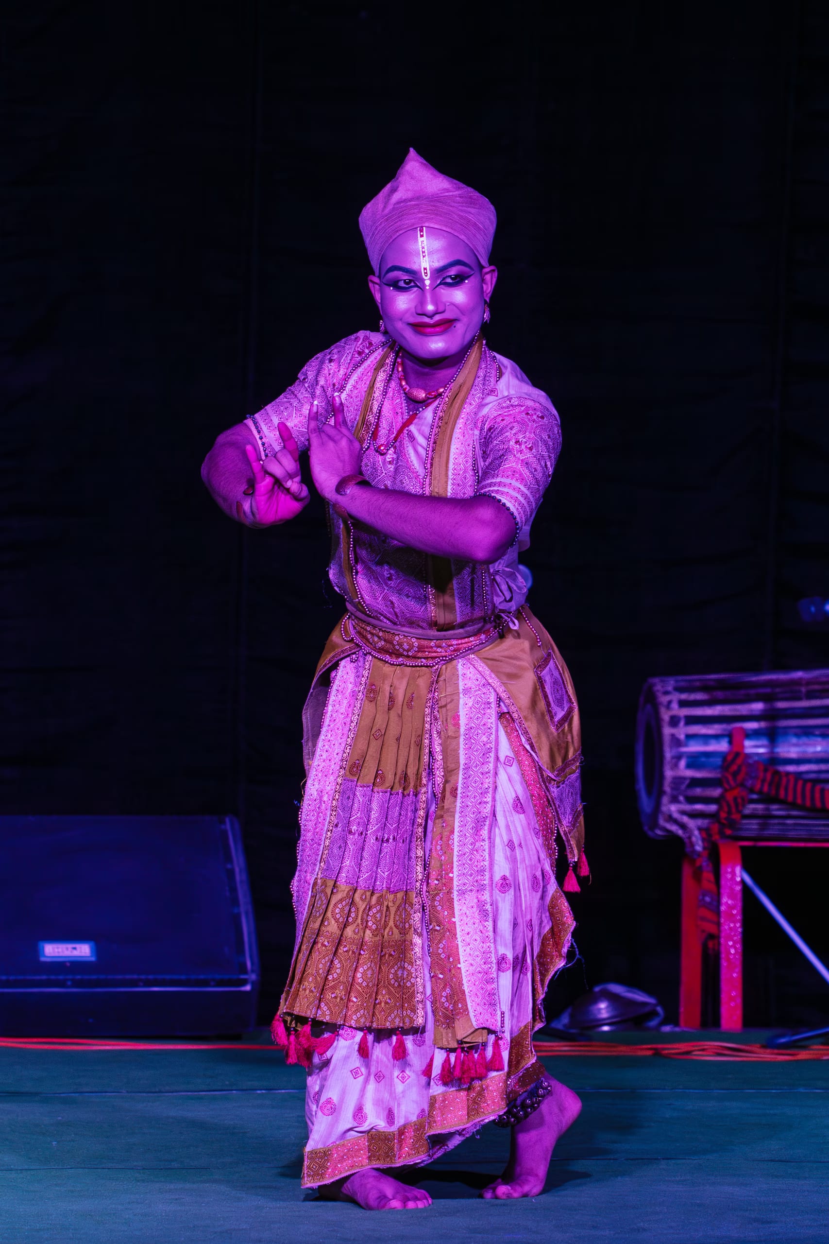 A Sattriya dancer in costume performing on stage, with a khol drum beside