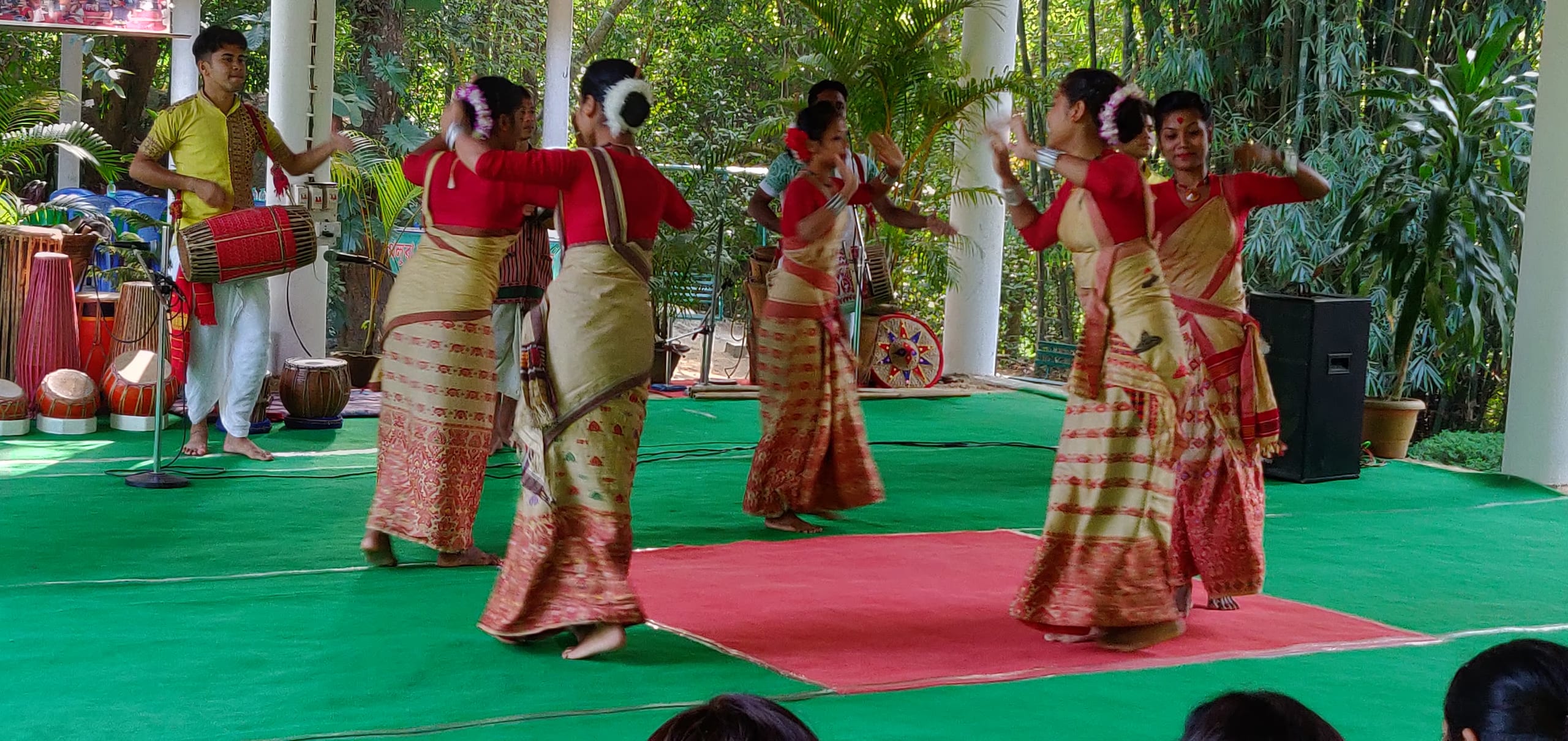 Women in red blouses and golden mekhela-sela dancing the Bihu with a dhol drummer at a stage