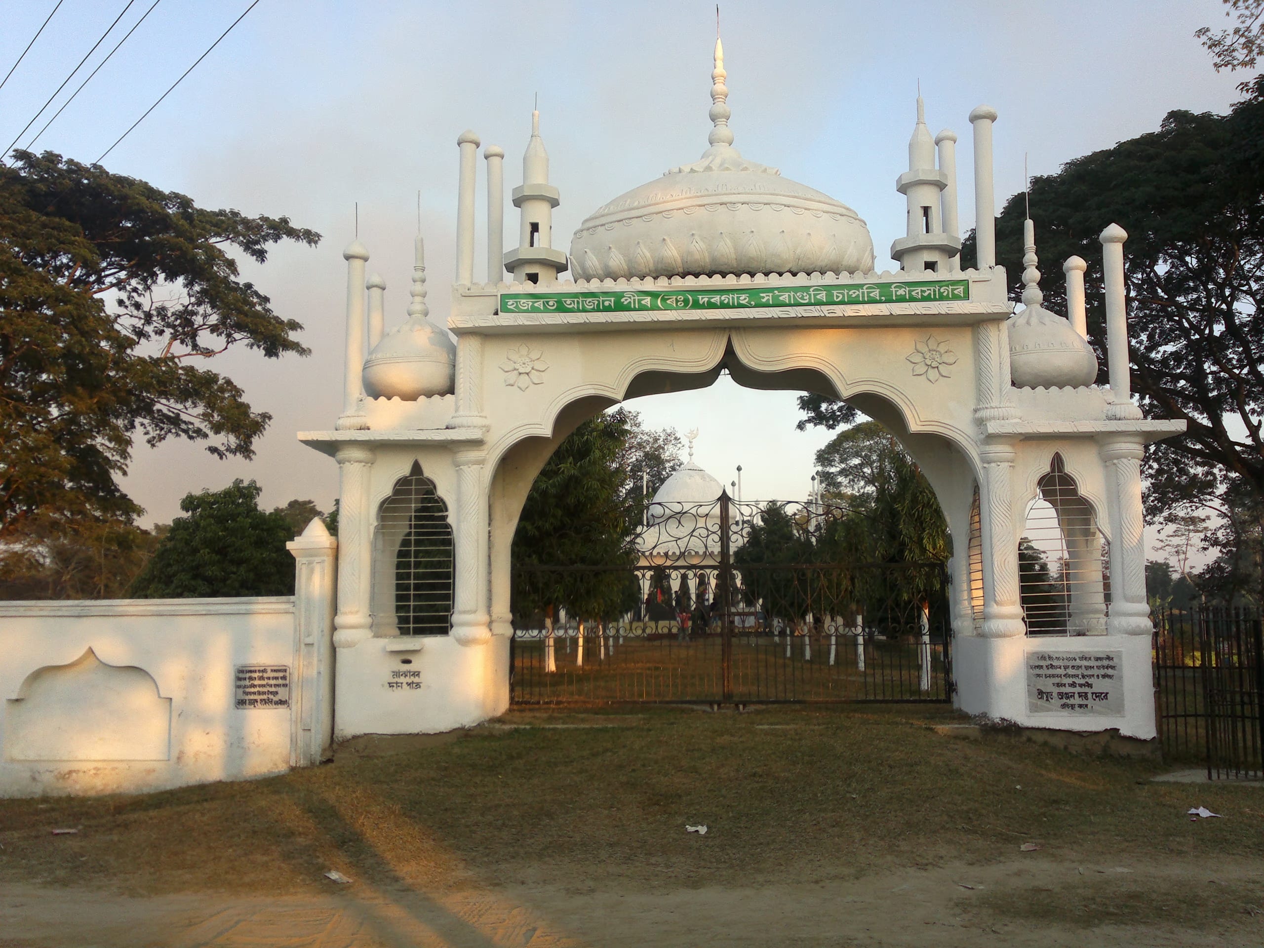 White domed gateway of the Ajan Pir Dargah with an Assamese signboard, the shrine visible through the archway beyond