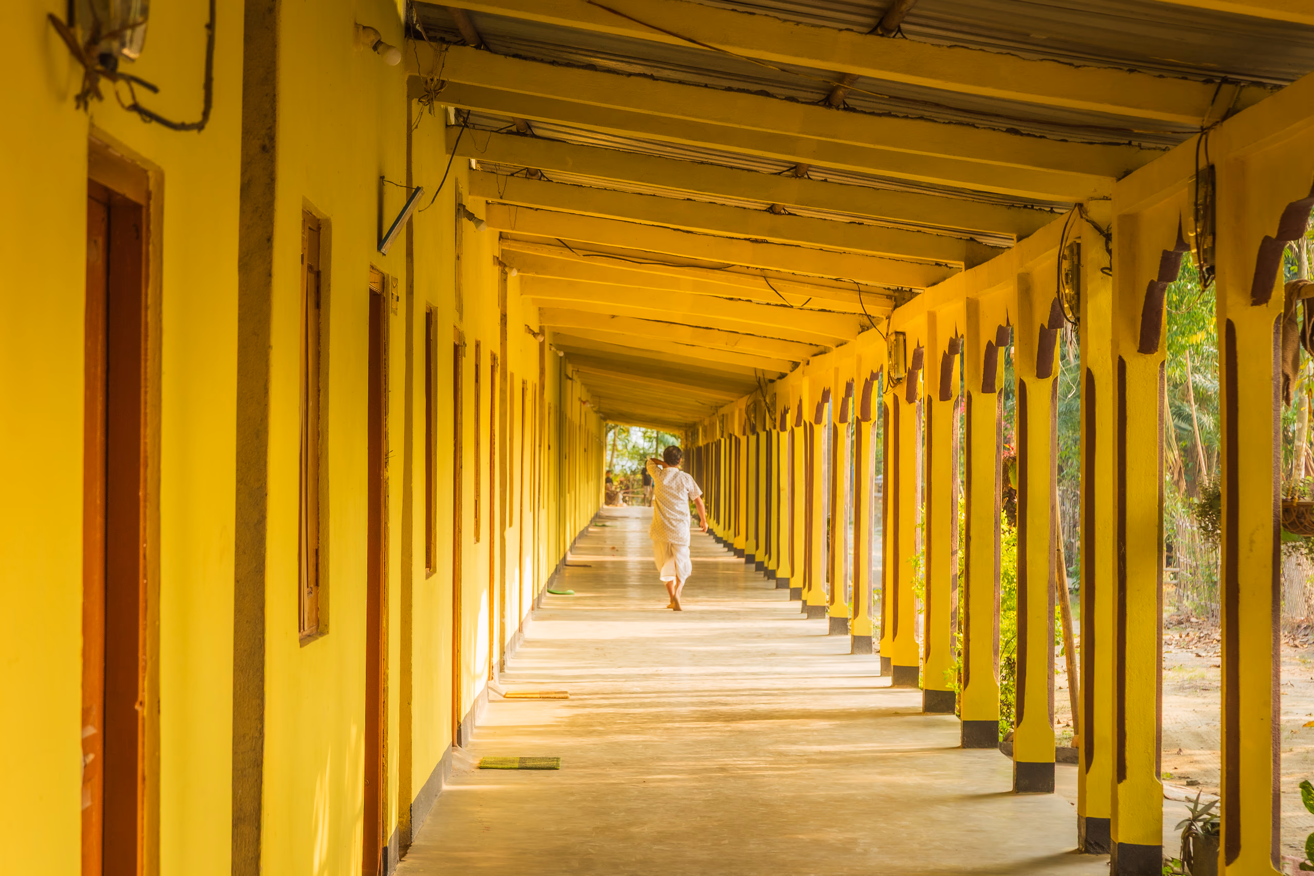 A long colonnade of yellow square pillars at Auniati Sattra receding to a bright vanishing point, a single distant figure walking down the shaded walkway