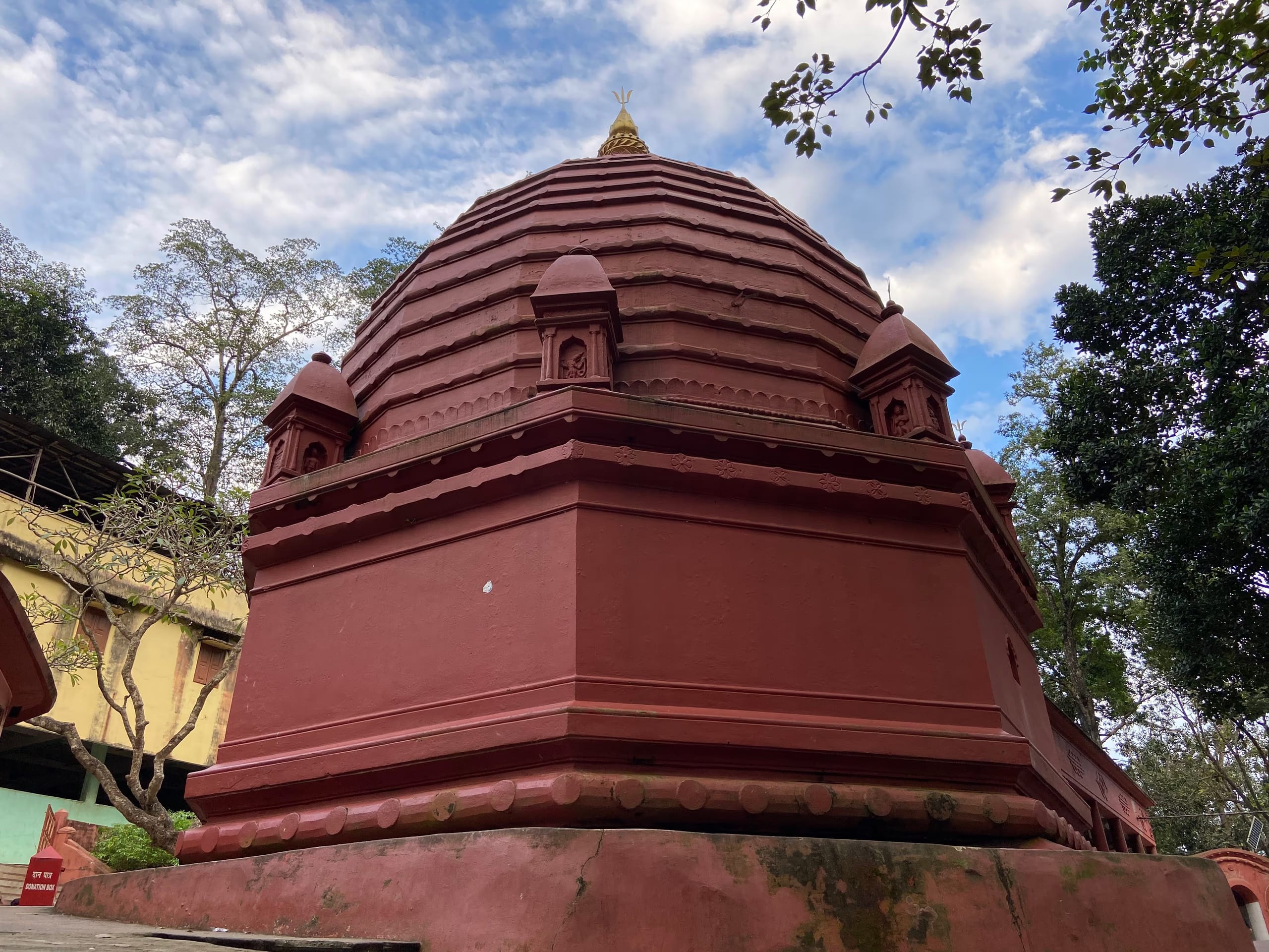The red-painted octagonal Siva temple at Basistha, its ribbed polygonal sikhara crowned by a gold finial, framed by trees against a blue sky