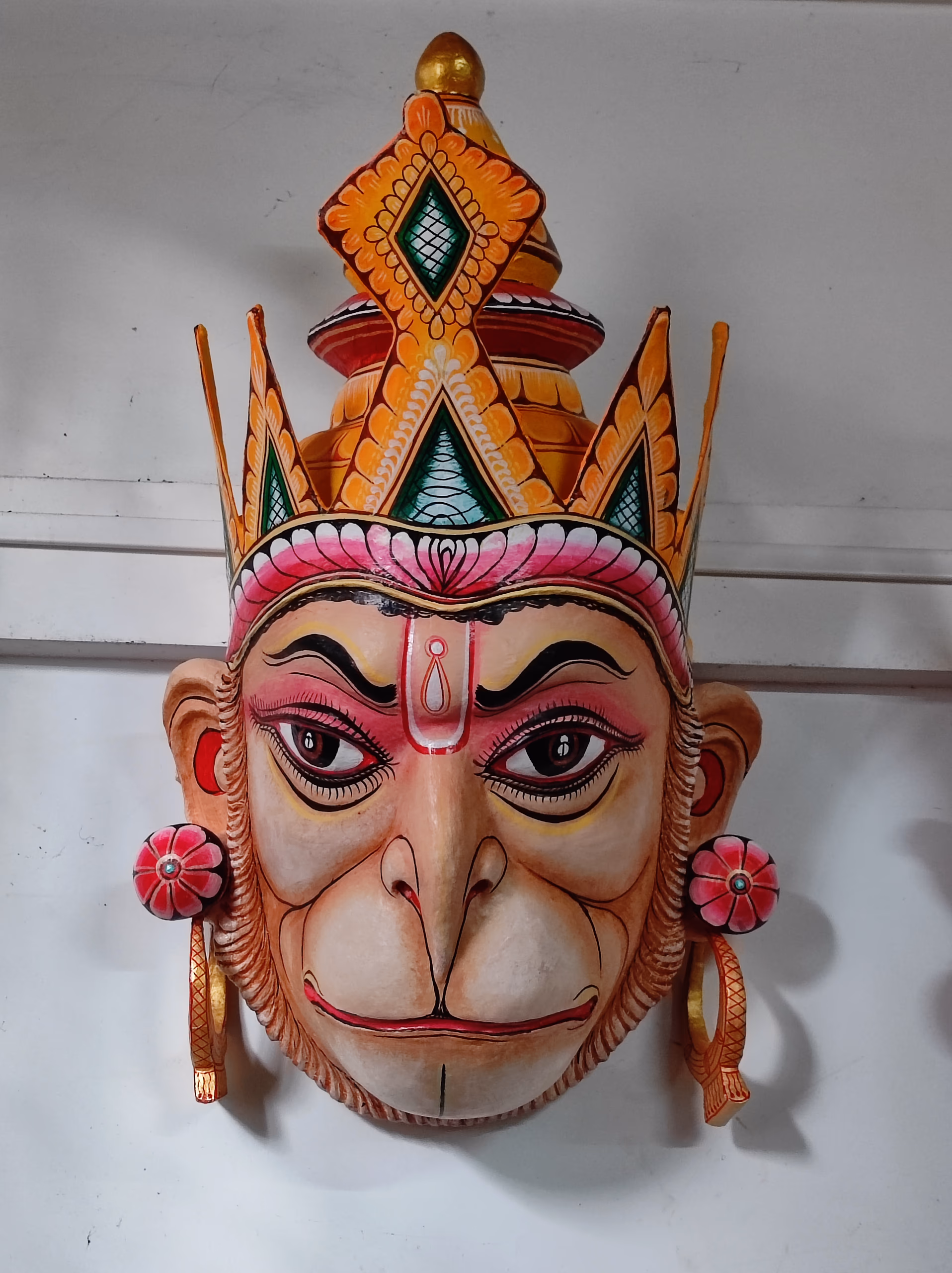 A brightly painted papier-mache mask of Hanuman with a crowned headdress, displayed against a plain white wall