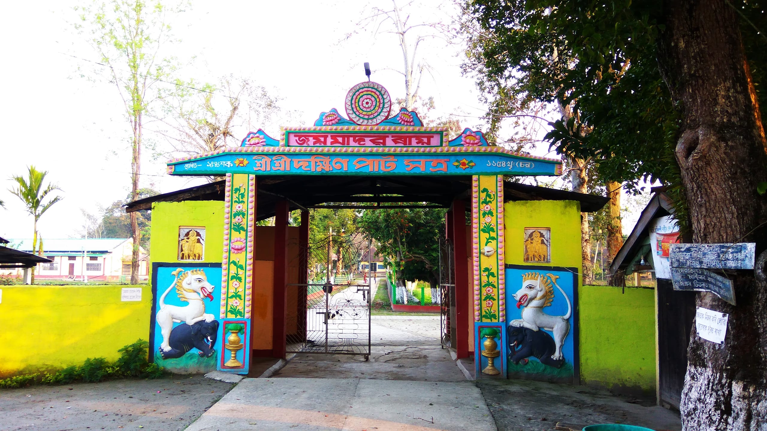A brightly painted gateway with a pitched roof, flanked by murals of a white horse and a lion, lettered in Assamese, opening onto the sattra grounds