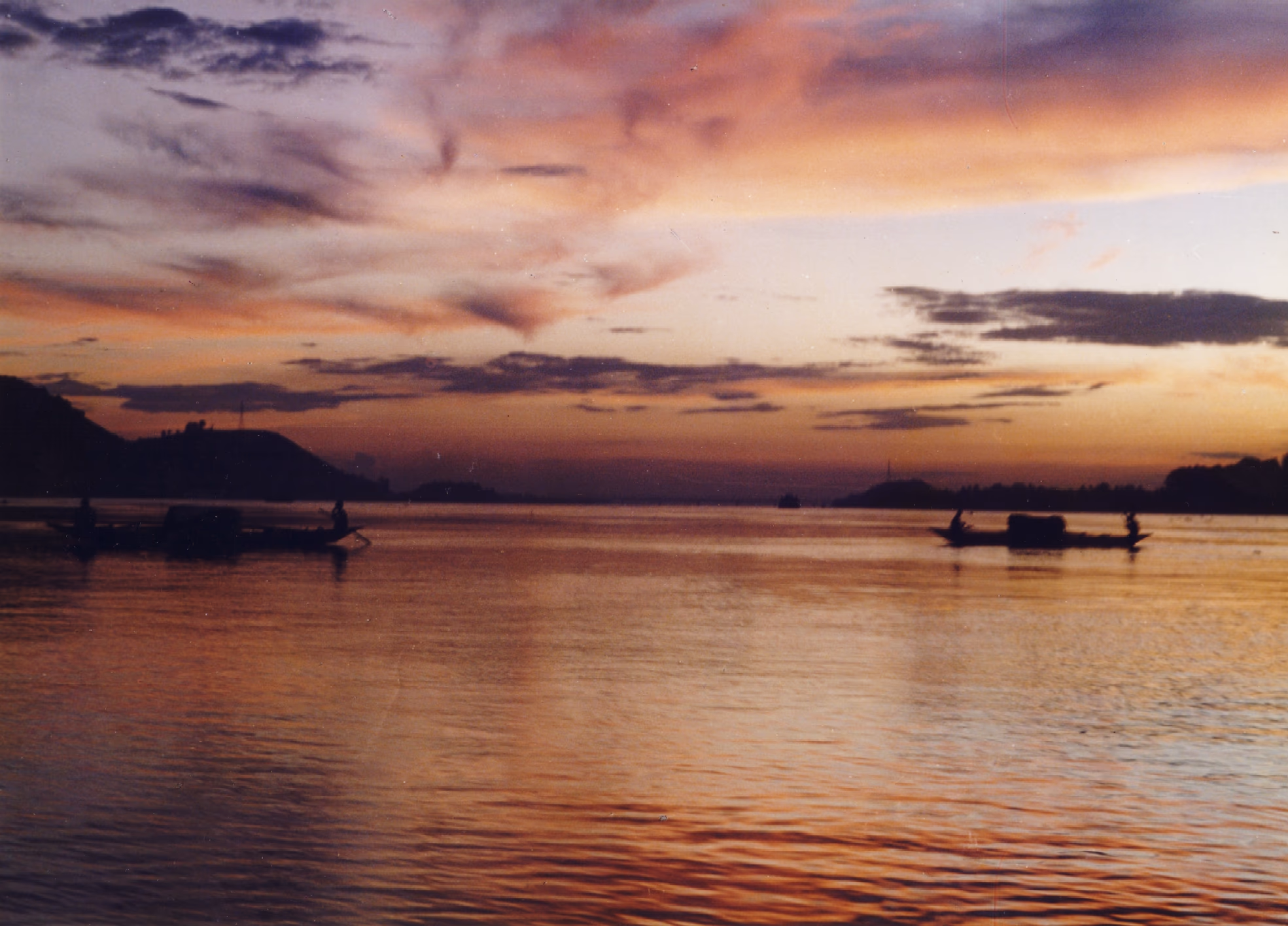 Wooden boats with standing rowers silhouetted on the broad, calm Brahmaputra at sunset off Guwahati, the river burning pink and gold under a dramatic sky with low hills along both banks