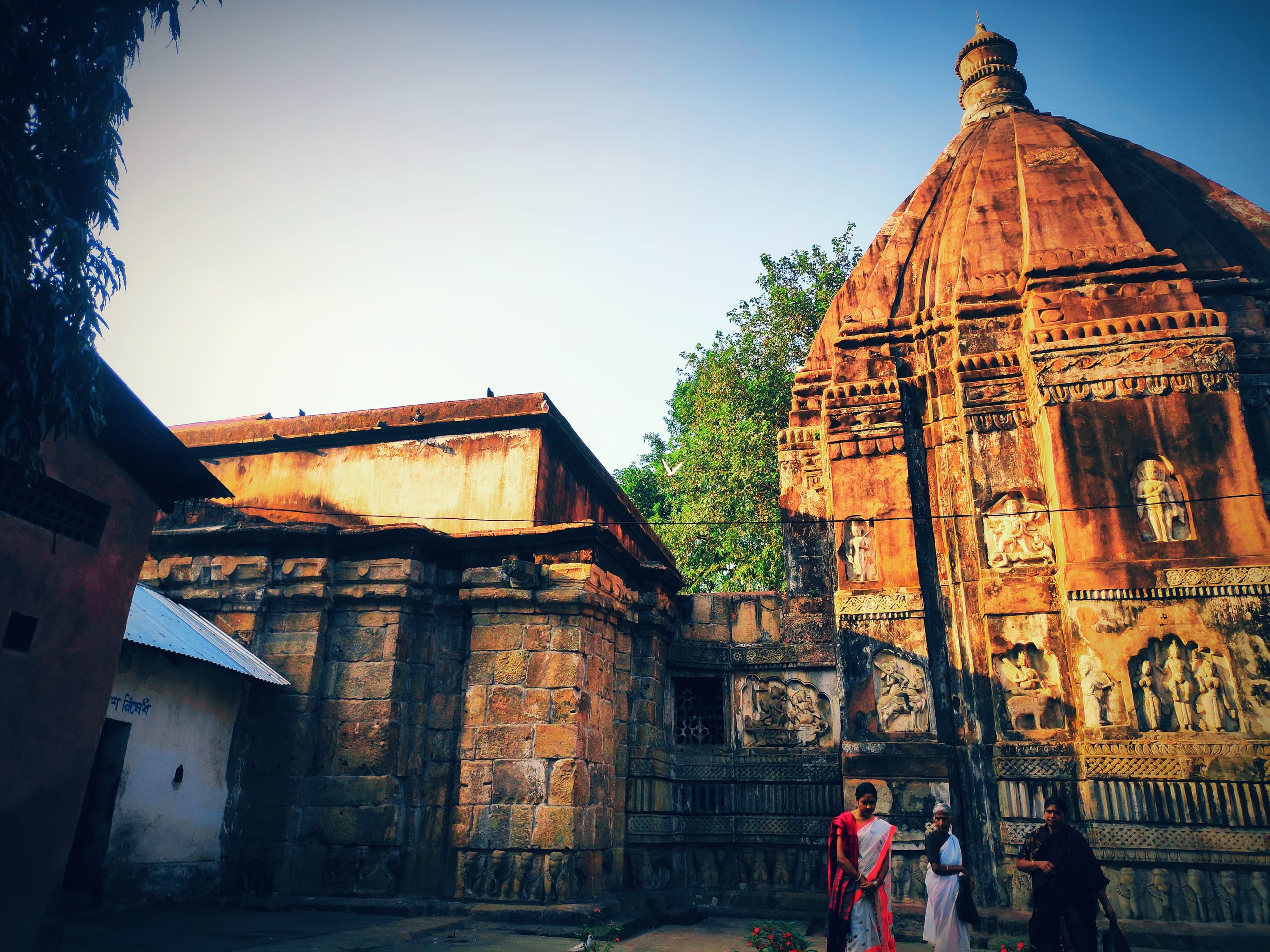 The laterite-red stone Hayagriva Madhava temple at Hajo in warm evening light, its curved carved shikhara tower beside the lower hall, framed by green trees under a deep blue sky