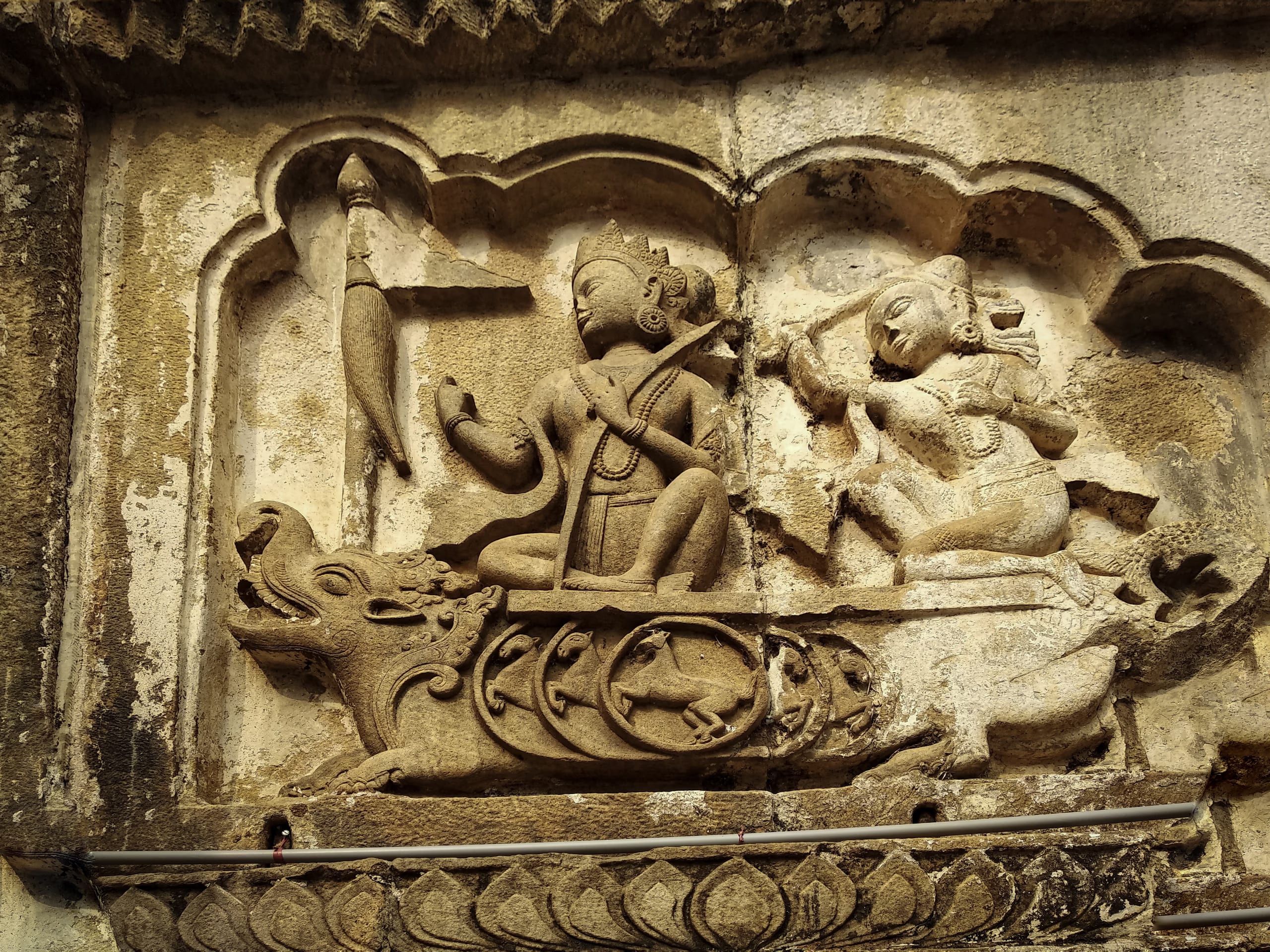 A sandstone relief panel set in a cusped niche on the temple wall: a crowned figure riding a makara, a horned aquatic beast, and holding a stringed instrument, faced by a second seated figure, above a band of carved lotus-petal ornament