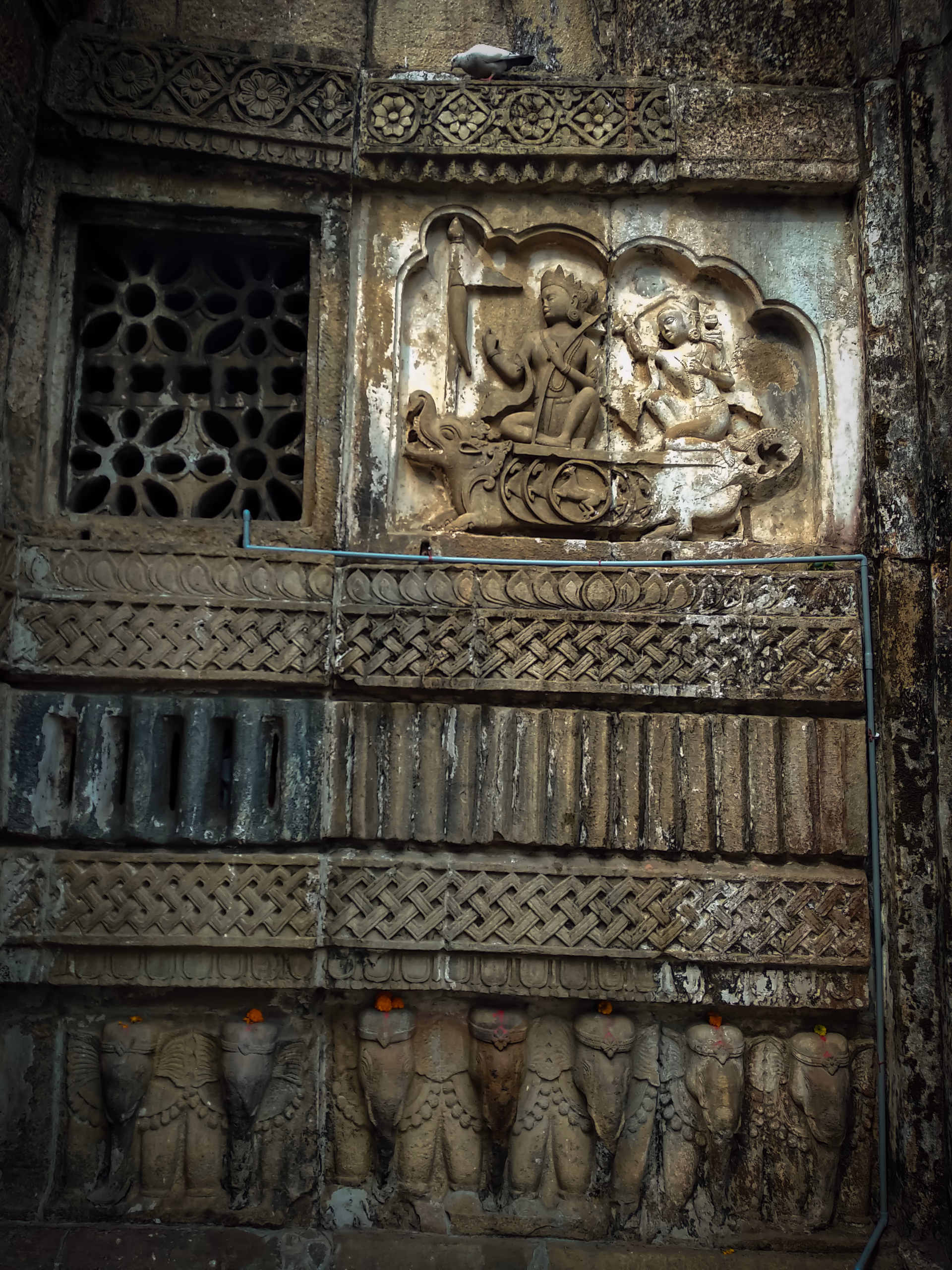 A weathered stone temple wall: a sandstone relief of a deity in a boat set in a cusped niche, beside a pierced lattice (jali) window, above carved ornamental bands and a base course of elephant-head sculptures hung with marigold garlands