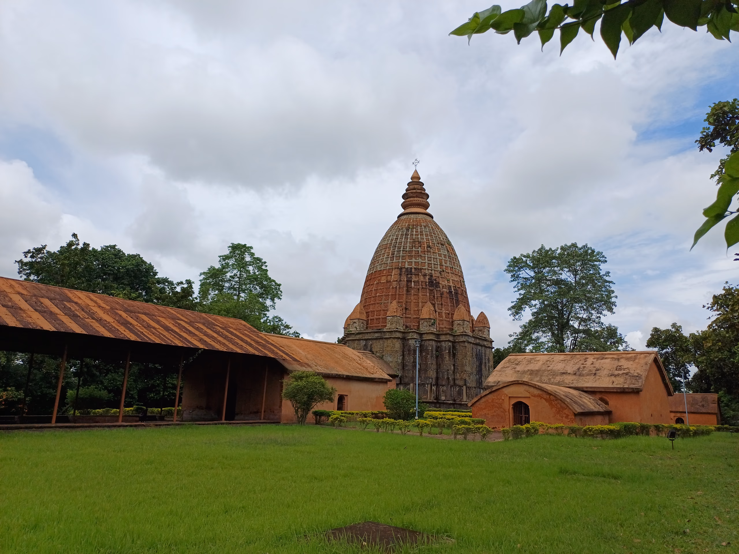 Low-angle view across a green lawn to the brick Joydol at Joysagar: a ribbed curvilinear Ahom dol tower crowned by a tiered amalaka and trident finial over a square carved sanctum, with an open pillared hall to the left and a barrel-roofed brick shrine to the right, under a dramatic monsoon sky