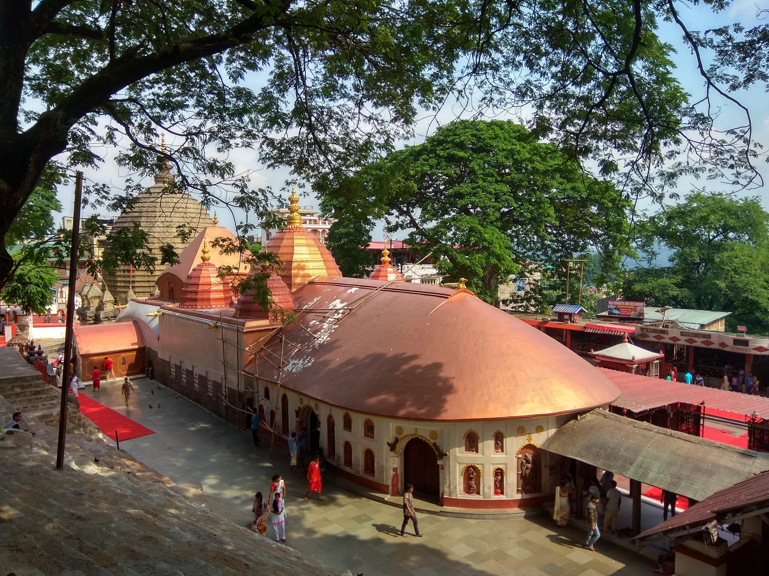 Elevated daytime view of the Kamakhya temple complex: the curved copper-coloured roof of the natamandira in front, the beehive shikhara and tiered red turrets rising behind, framed by green trees