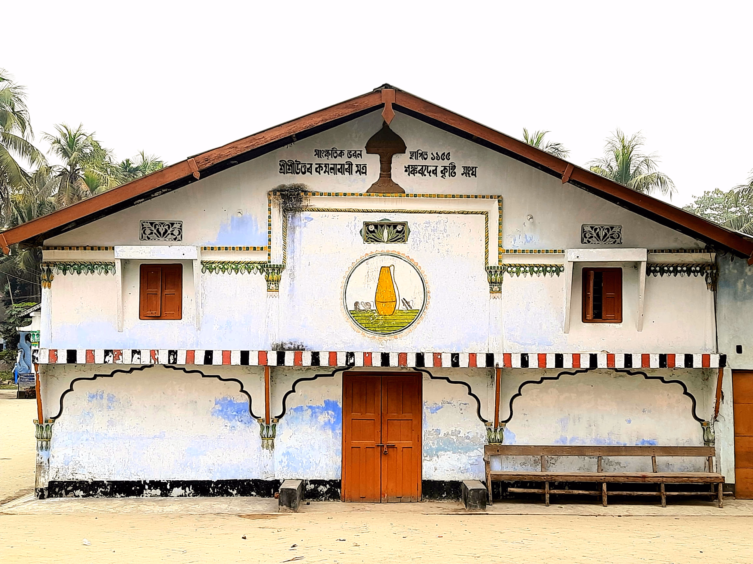 Whitewashed prayer-hall facade of the Uttar Kamalabari Sattra on Majuli, with an Assamese inscription in the gable, a central doorway and a painted emblem