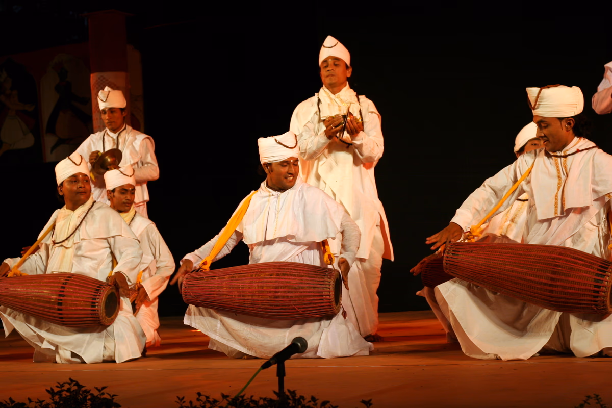 Men in white dress and white turbans kneeling and playing the khol drum, with a cymbal player standing, on a performance stage