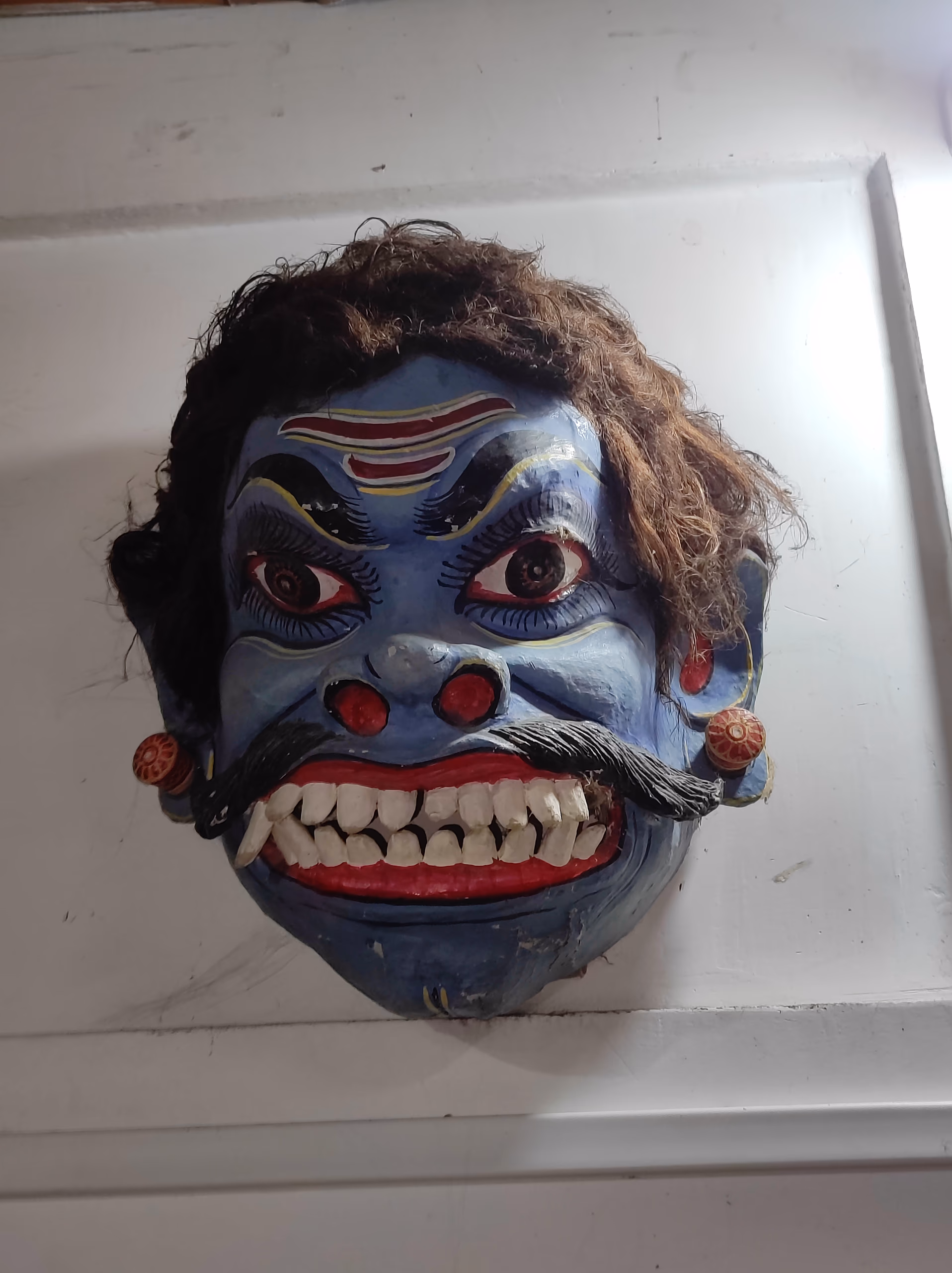 Blue-faced demon mukha with bulging eyes, bared white teeth, a thick black moustache and dark hair, a Majuli mask for a bhaona villain