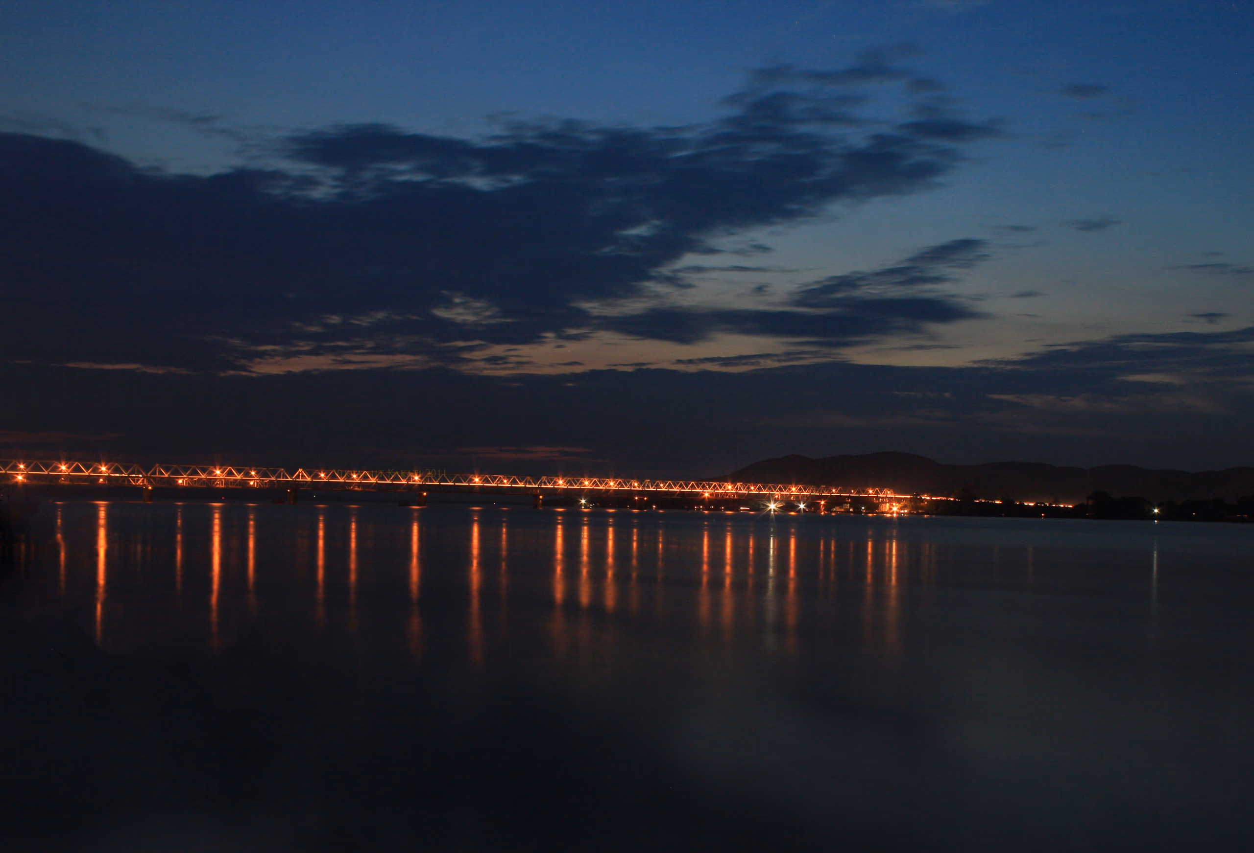 The Saraighat road-and-rail bridge, its truss span lit and reflected on the Brahmaputra at dusk, with low hills on the far bank