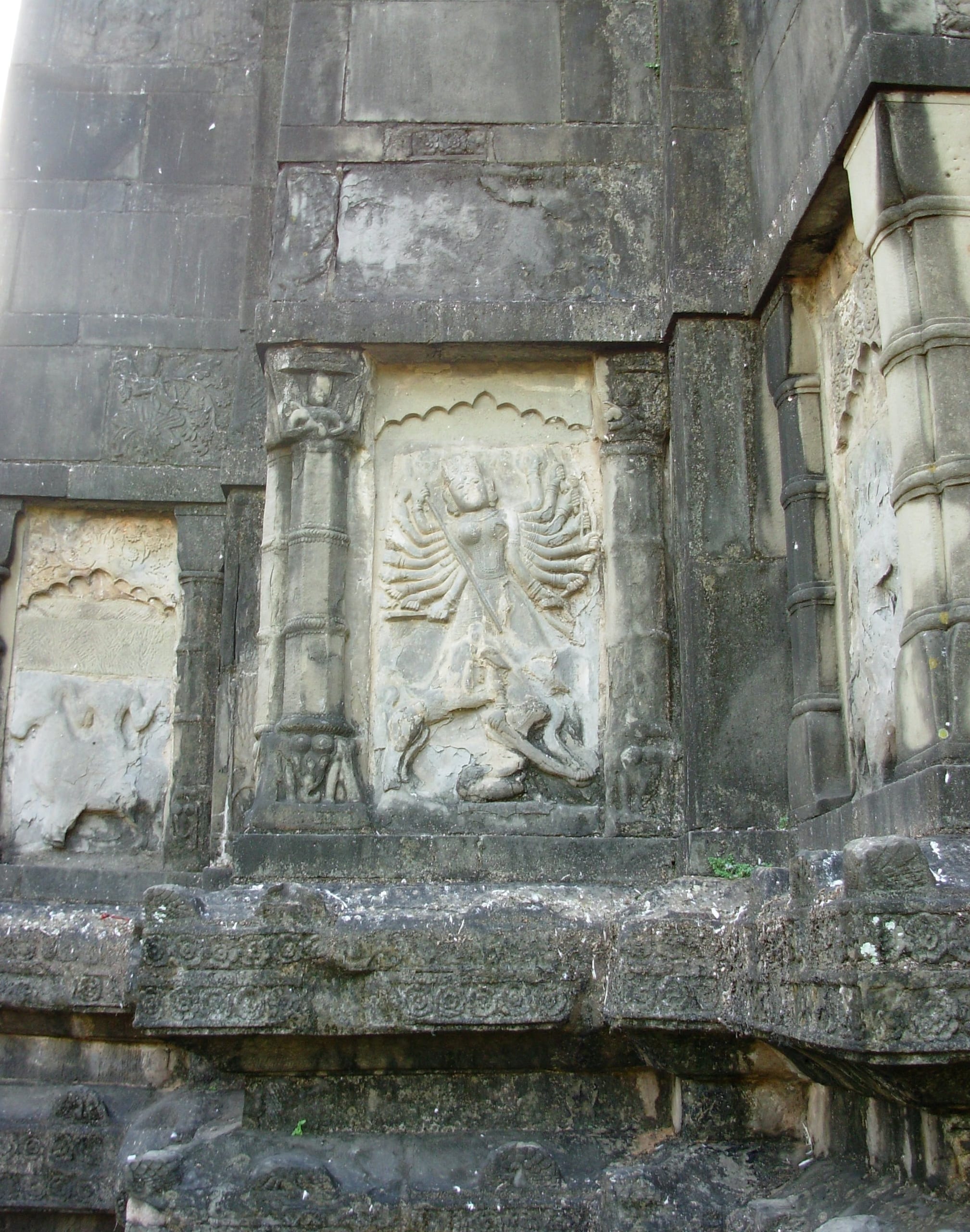 Carved stone relief in a cusped wall niche on the dark stone temple wall, showing a many-armed goddess astride and slaying a figure below, flanked by carved colonnettes above a decorated frieze band