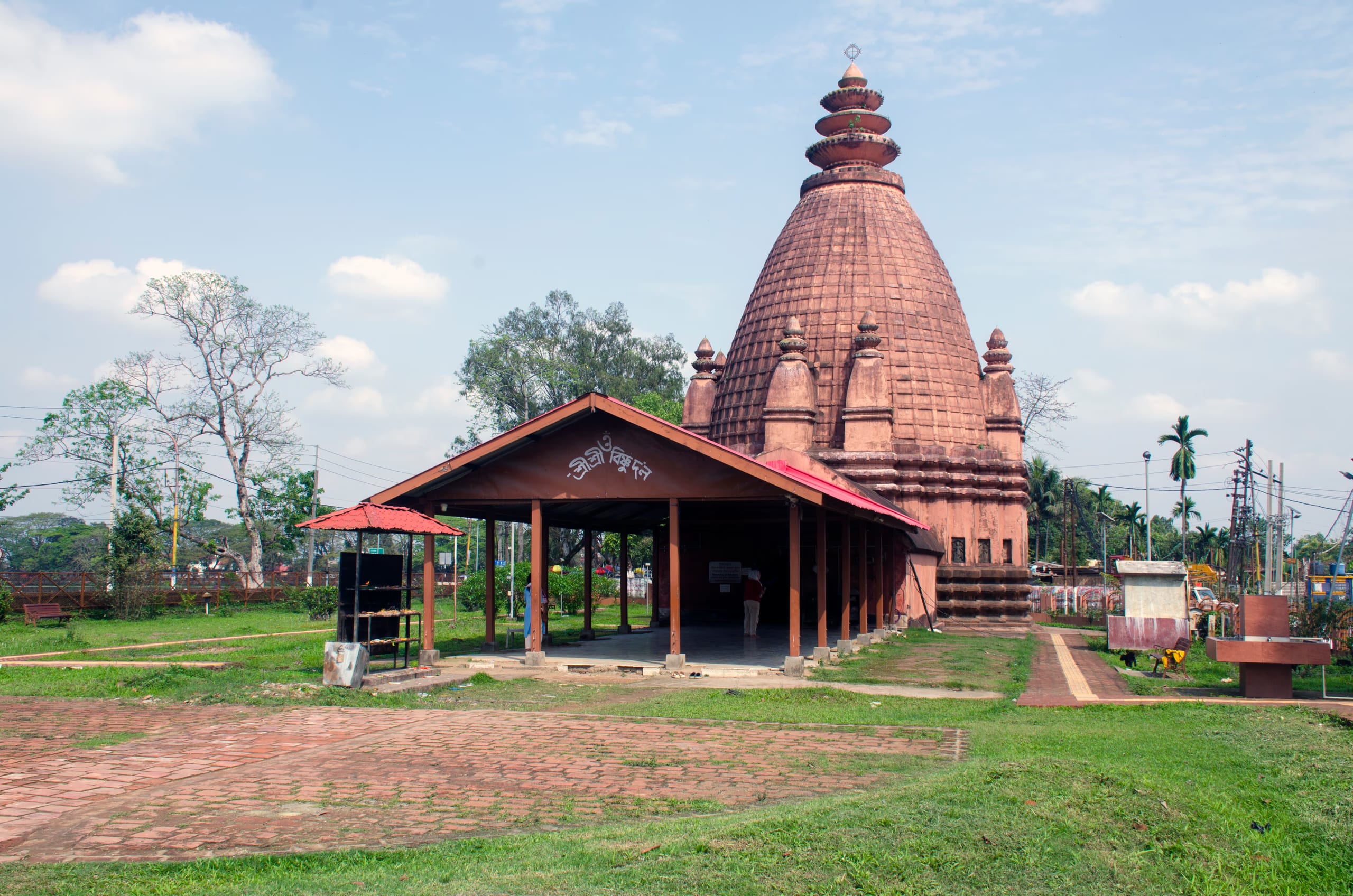 A red-brick Ahom temple with a tall ribbed curvilinear shikhara and a low open-sided mandapa pavilion in front, standing on a brick-paved lawn under a pale blue sky
