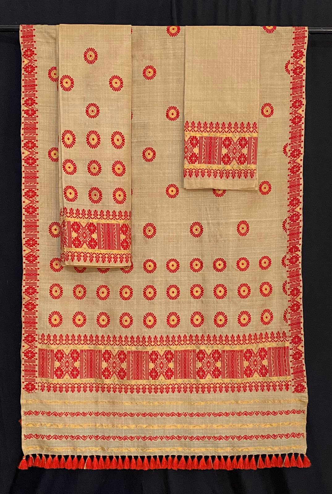 A two-piece muga silk mekhela-sador laid on a black ground, its natural golden cloth worked with rows of red sunflower-like medallions and broad red-and-gold patterned borders