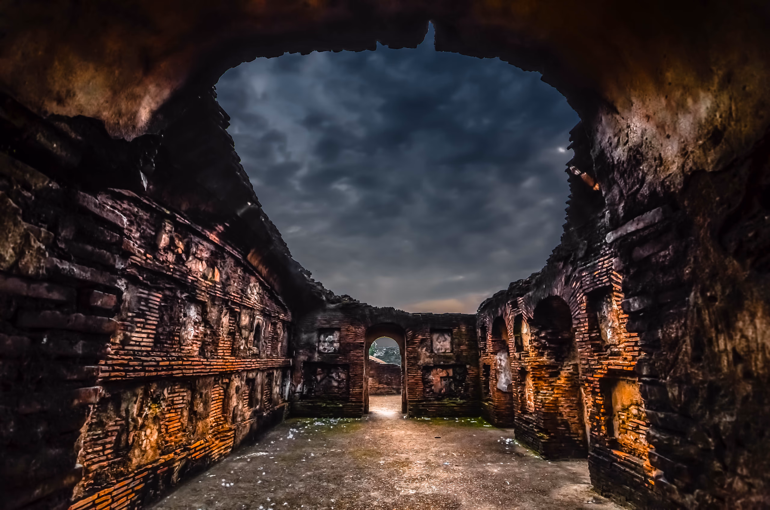 Inside a roofless brick chamber of the Talatal Ghar, the broken vault open to a dark cloudy sky, warm light raking the thin Ahom brick walls and a lit doorway at the far end