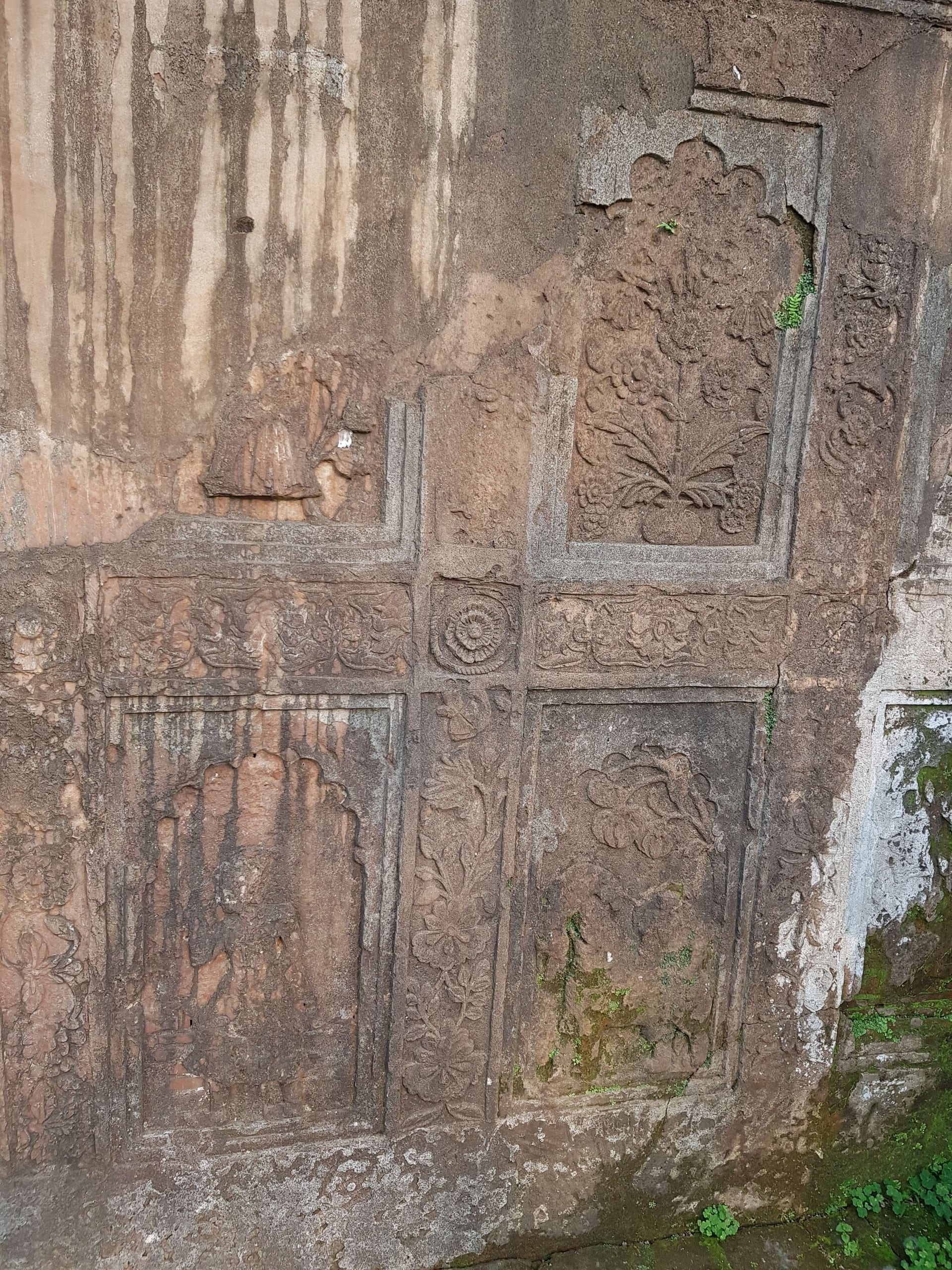 A weathered carved plaster-and-brick relief panel inside the Talatal Ghar, a cross-shaped frame of floral scrollwork enclosing an arched niche with a flowering-plant motif