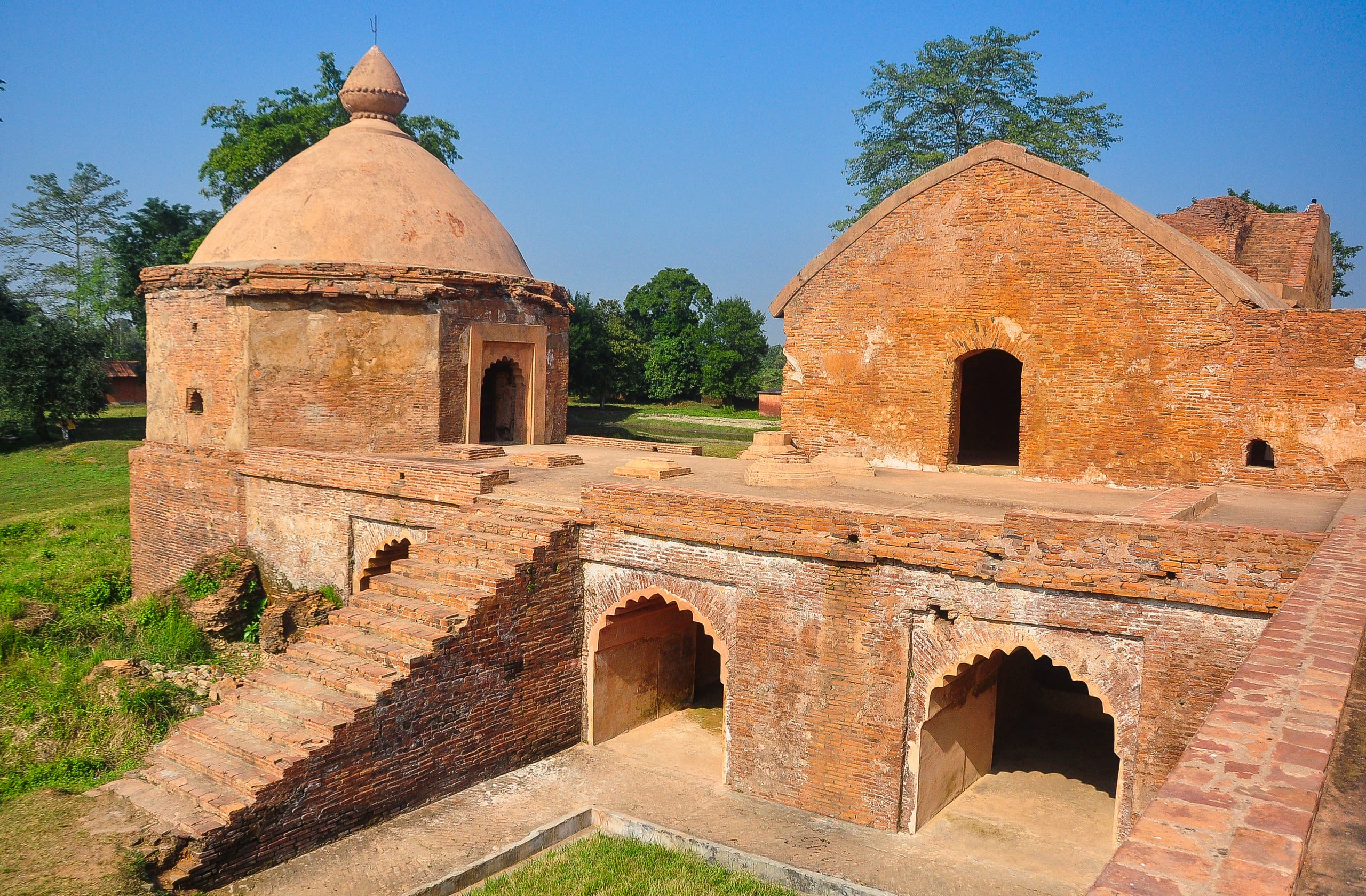 Weathered brick ruins of the Talatal Ghar with a domed pavilion, a gabled hall and several cusped arched openings under a clear sky