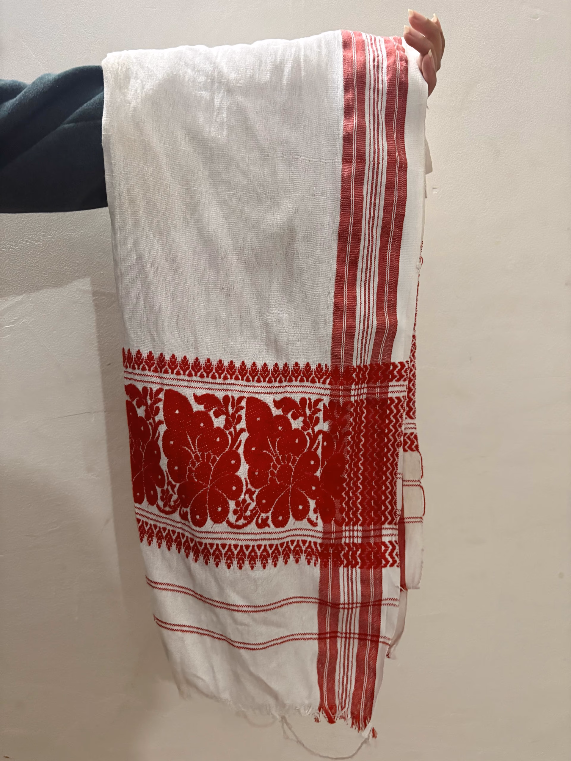 A handwoven white cotton gamosa with red end-borders and a broad band of red woven floral motifs, held up against a pale wall