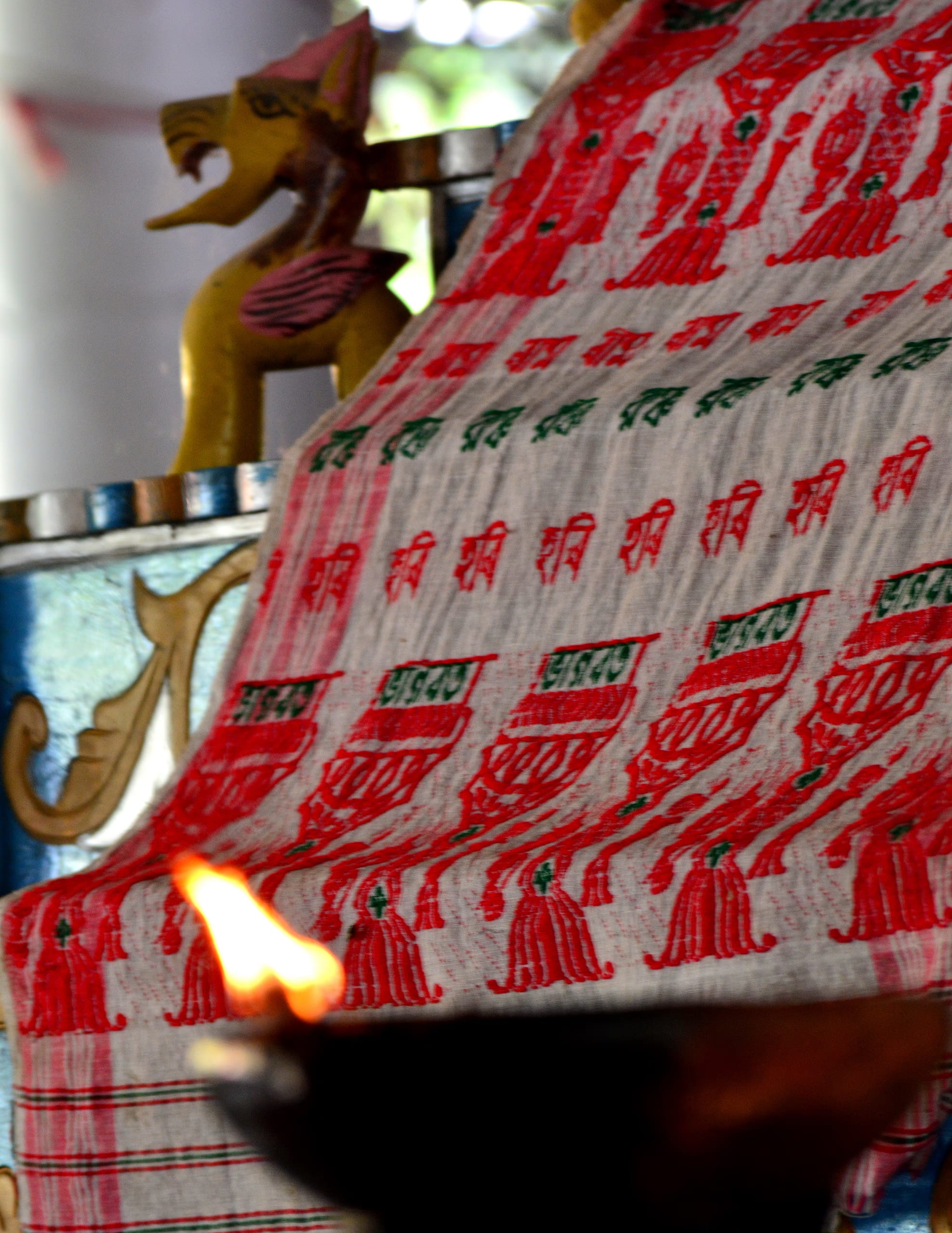 A patterned gamosa draped over a shrine offering-stand, a small oil lamp burning before it and a carved wooden lion behind