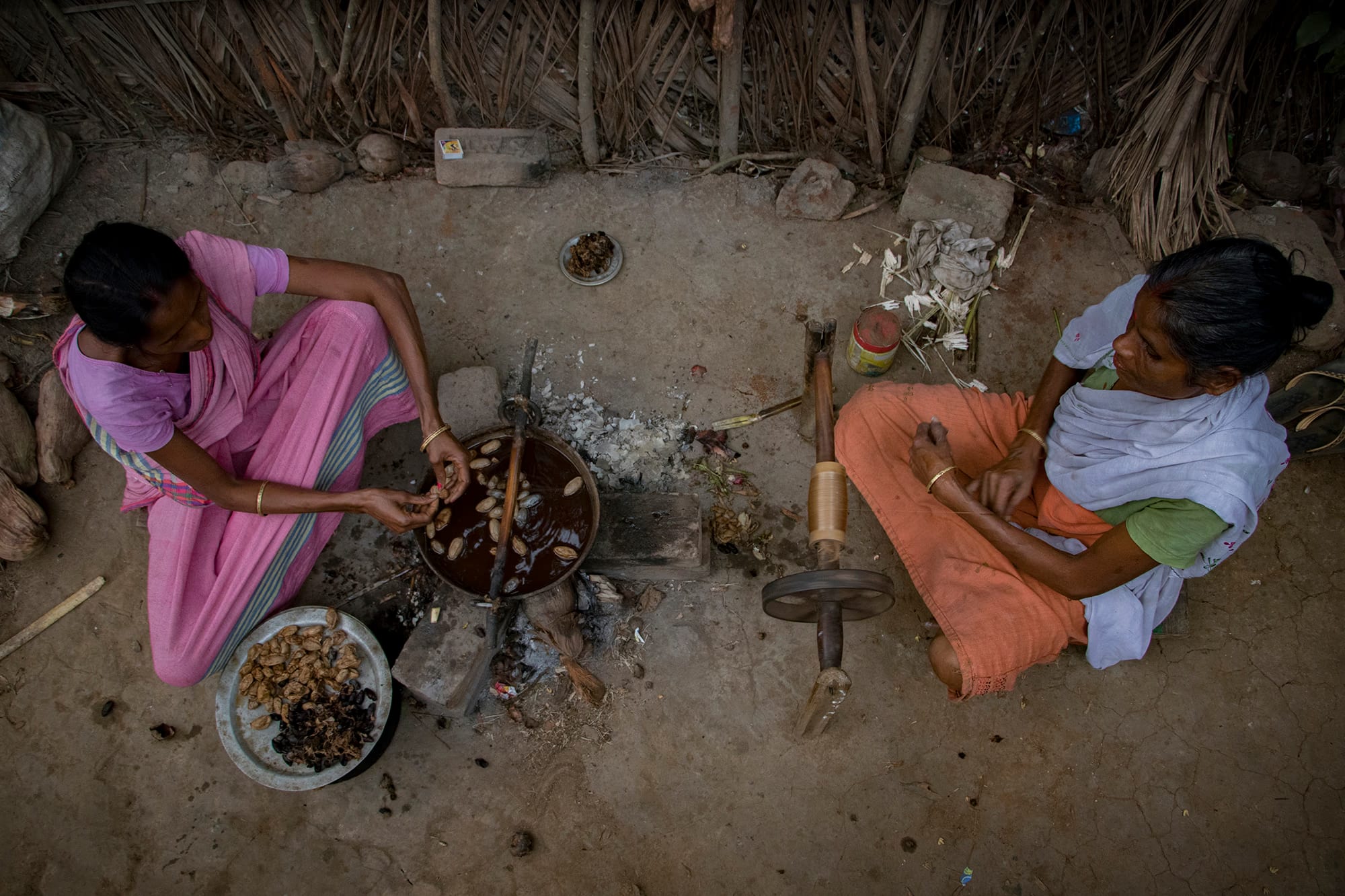 Two women reel muga silk, one softening cocoons in a pot, the other turning a hand spindle