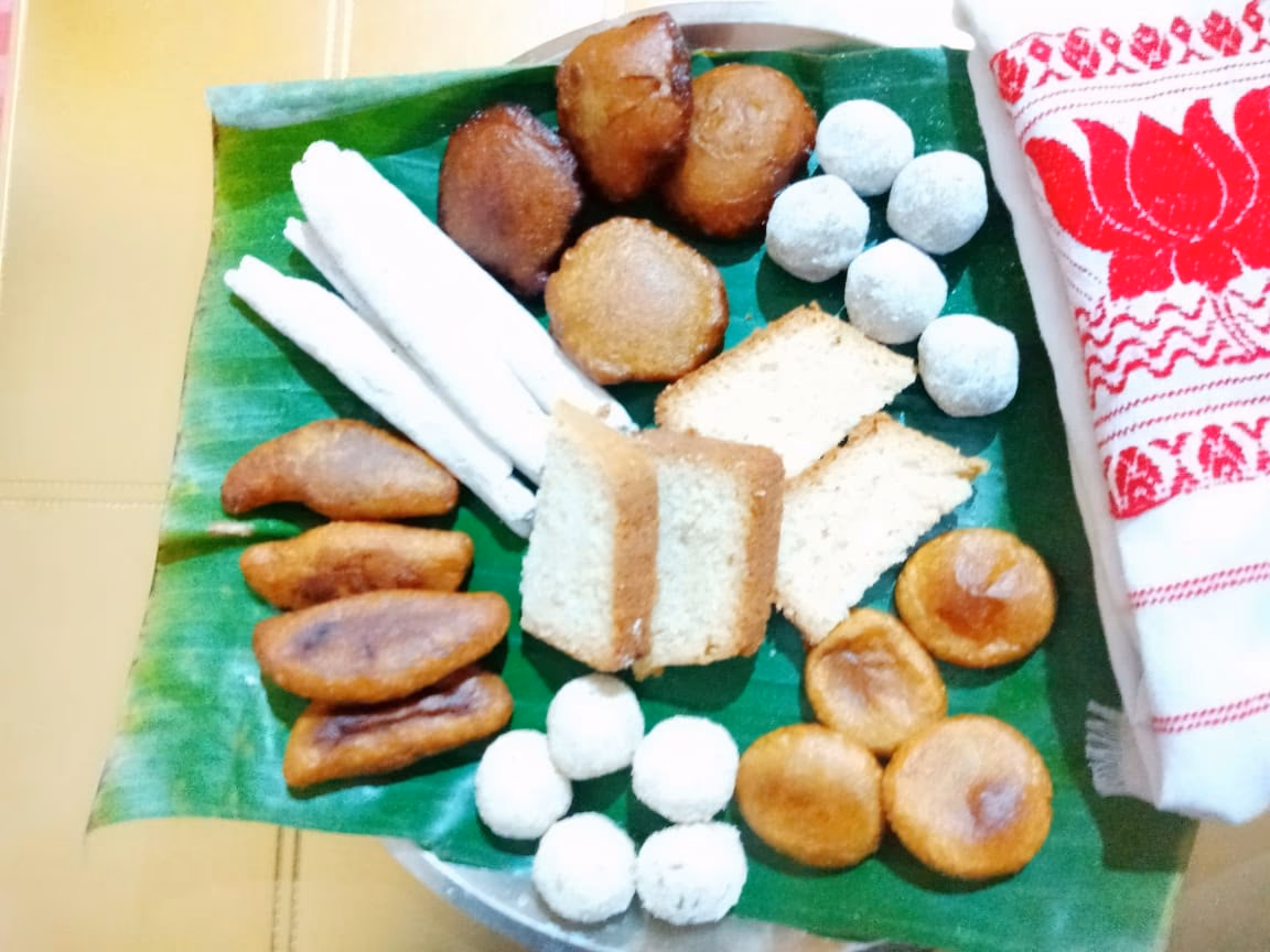 A banana-leaf platter of til pitha, fried tel pitha, golden ghila pitha and white coconut laru beside a red-and-white gamosa