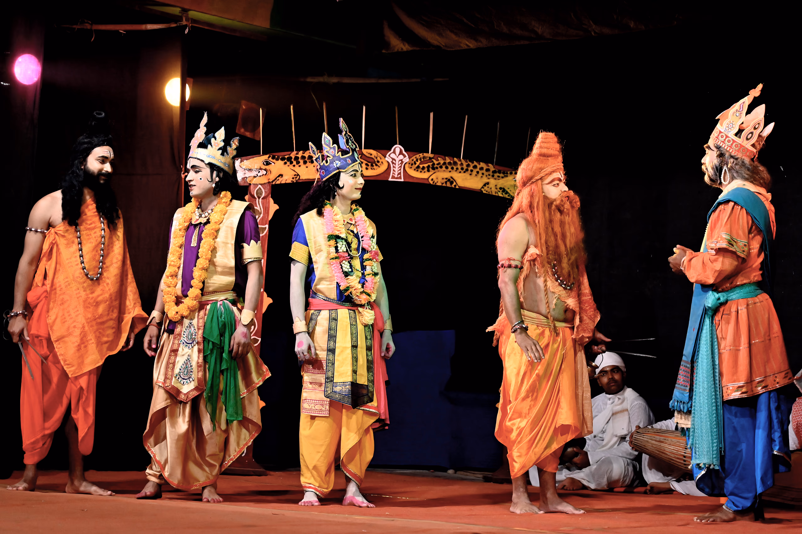 Costumed performers in robes, garlands and crowns stand on a sattra stage with a khol drummer seated behind them
