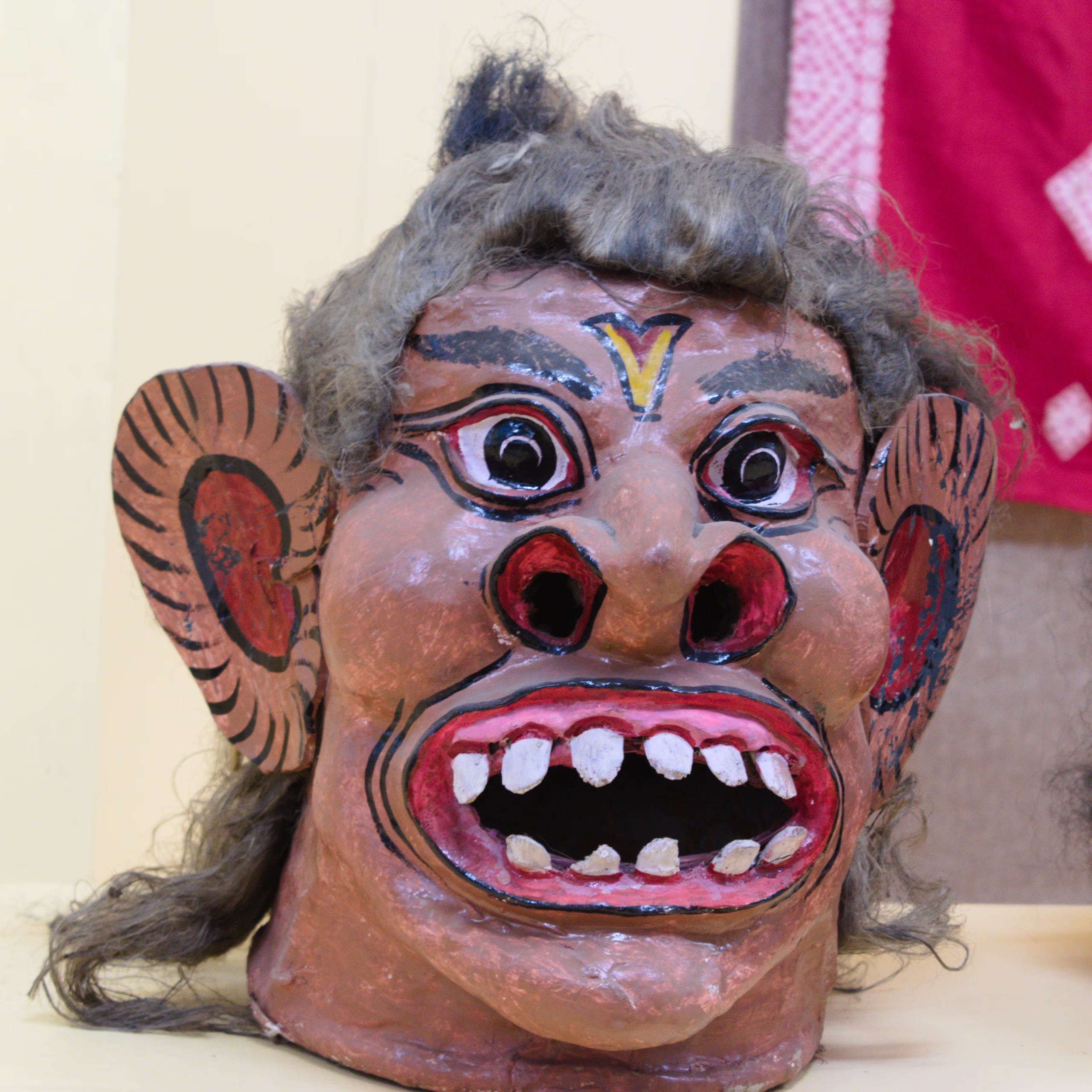 A painted papier-mache and clay mukha face-mask of a fierce demon, with a brown face, bulging red-rimmed eyes, a gaping red mouth showing white teeth, large painted side-ears and grey hair, displayed against a plain pale museum wall