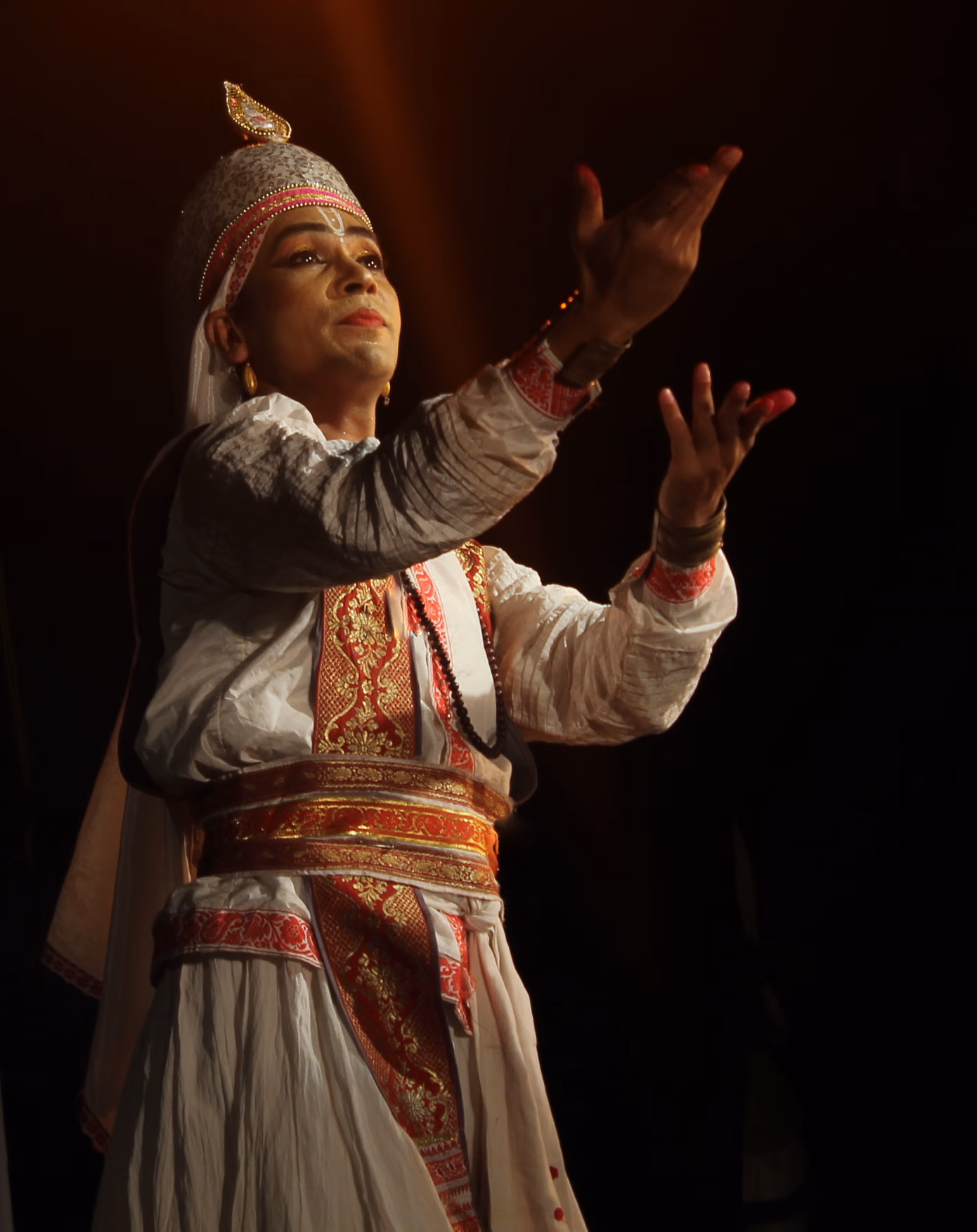 A solo performer in white-and-gold costume and a pointed crown raises both hands in a graceful gesture under a shaft of stage light against a dark background