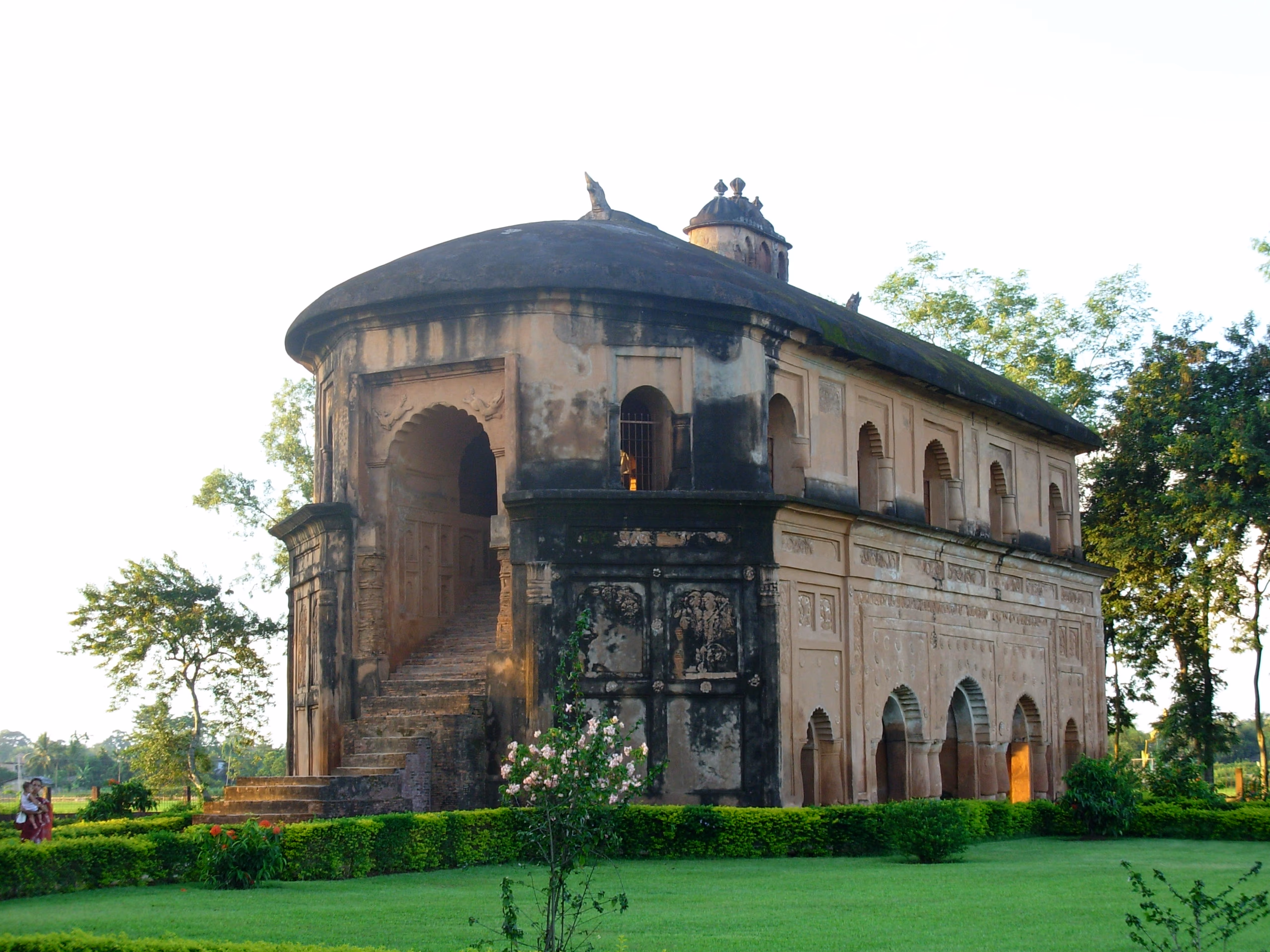 Rear three-quarter view of the Rang Ghar in warm afternoon light, showing the external masonry ramp that climbs to the upper gallery, with carved panels on the curved end and green surroundings