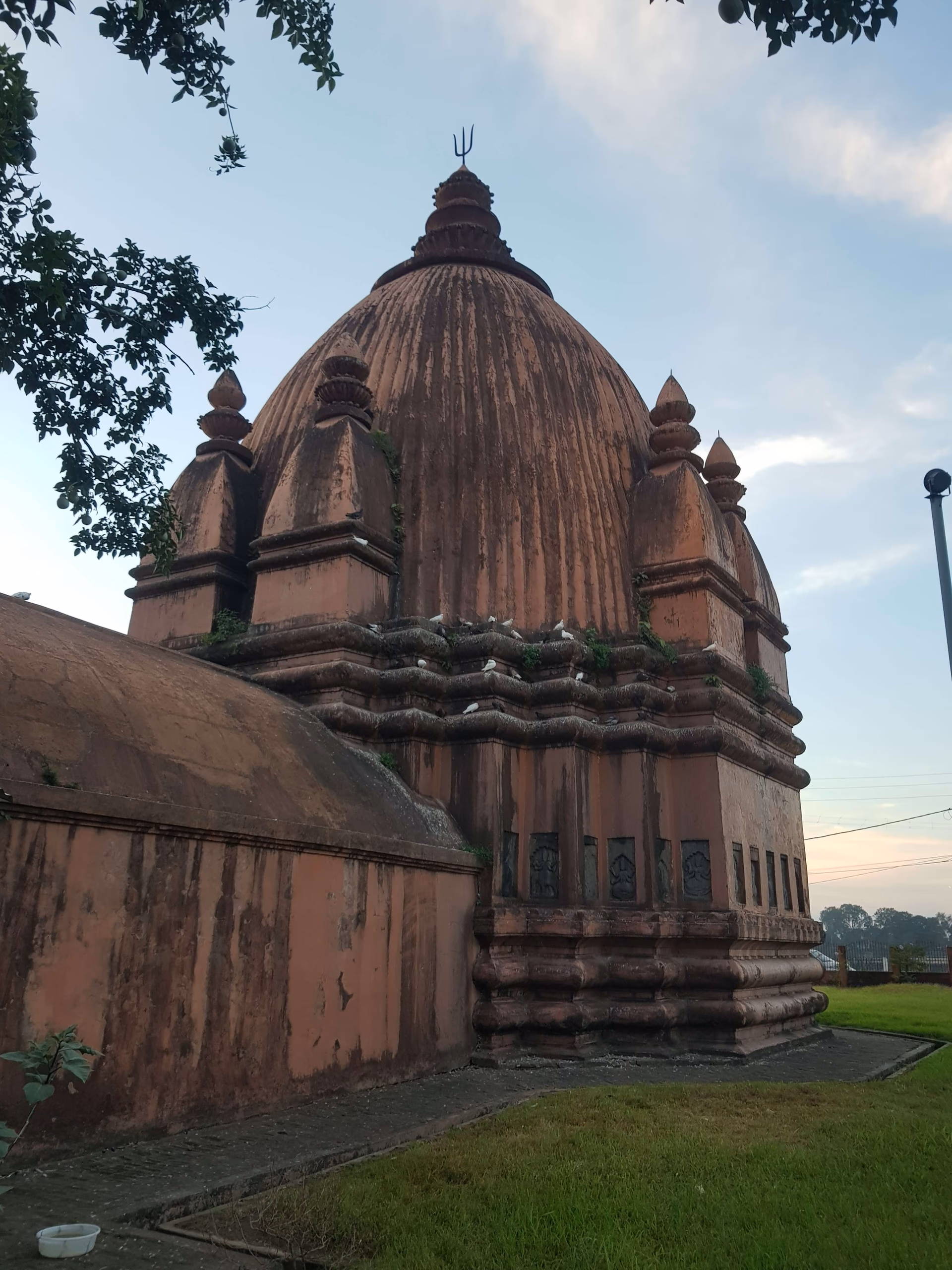 A red-brick Ahom temple seen three-quarter from below, its tall ribbed bell-shaped tower flanked by smaller corner turrets, pigeons perched on the cornices, against a soft pale-blue evening sky