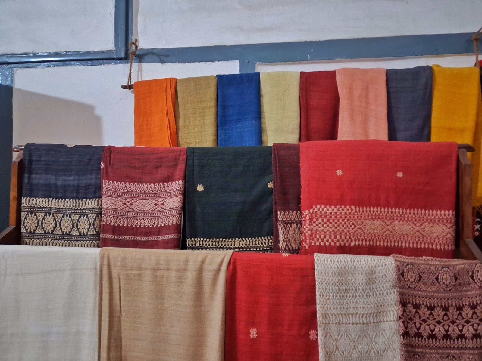 Rows of handwoven Assamese silk stoles and shawls hung on a wooden rack, in muga gold, eri cream and dyed red, blue, green and orange, each with woven floral and geometric border bands