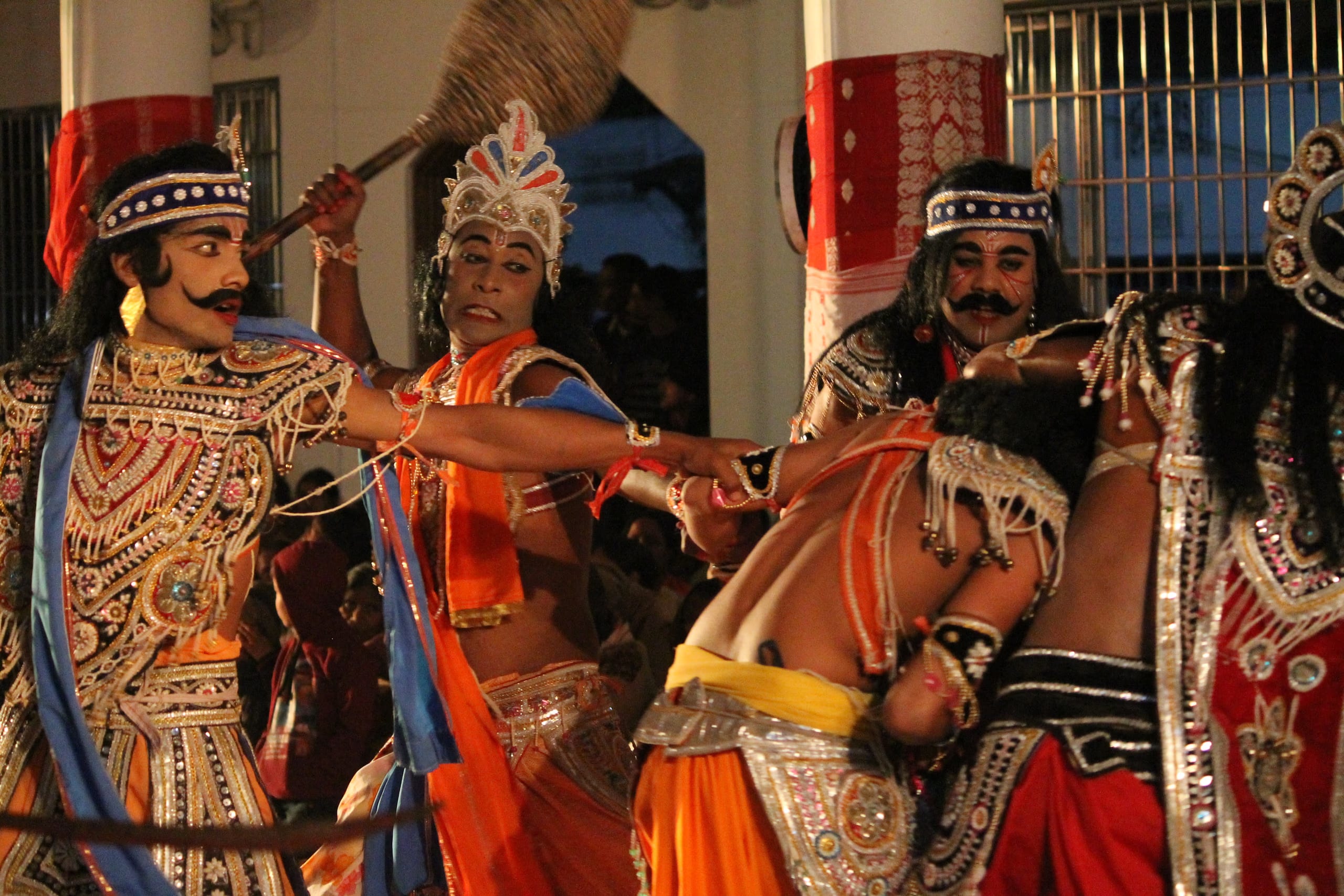 A bhaona stage scene at night: costumed actors in elaborate jewelled armour, crowns and painted faces clash mid-fight, one warrior raising a club, others grappling, all in spangled silver-and-gold dress with mirror-work, garlands and headdresses