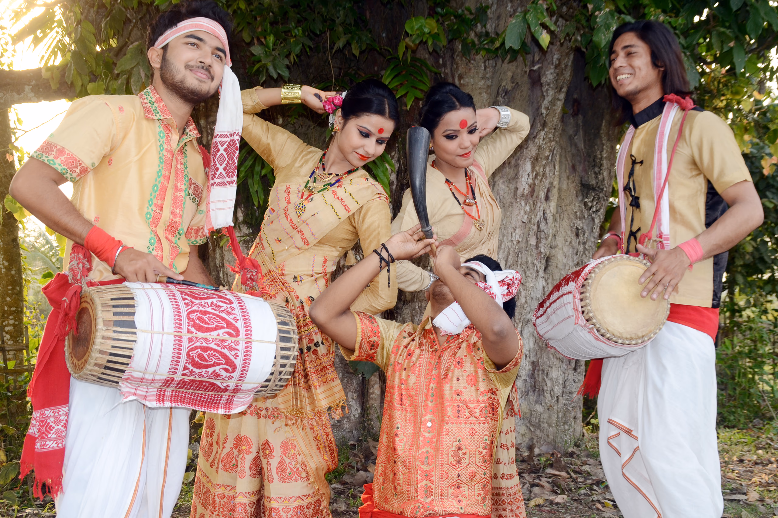 A posed group under a tree: two young men in cream kurtas and red-bordered gamosa each carrying a dhol slung at the waist, flanking two women in cream-and-red Bihu dress with hands raised to their hair, while a third man kneeling in front blows a buffalo-horn pepa